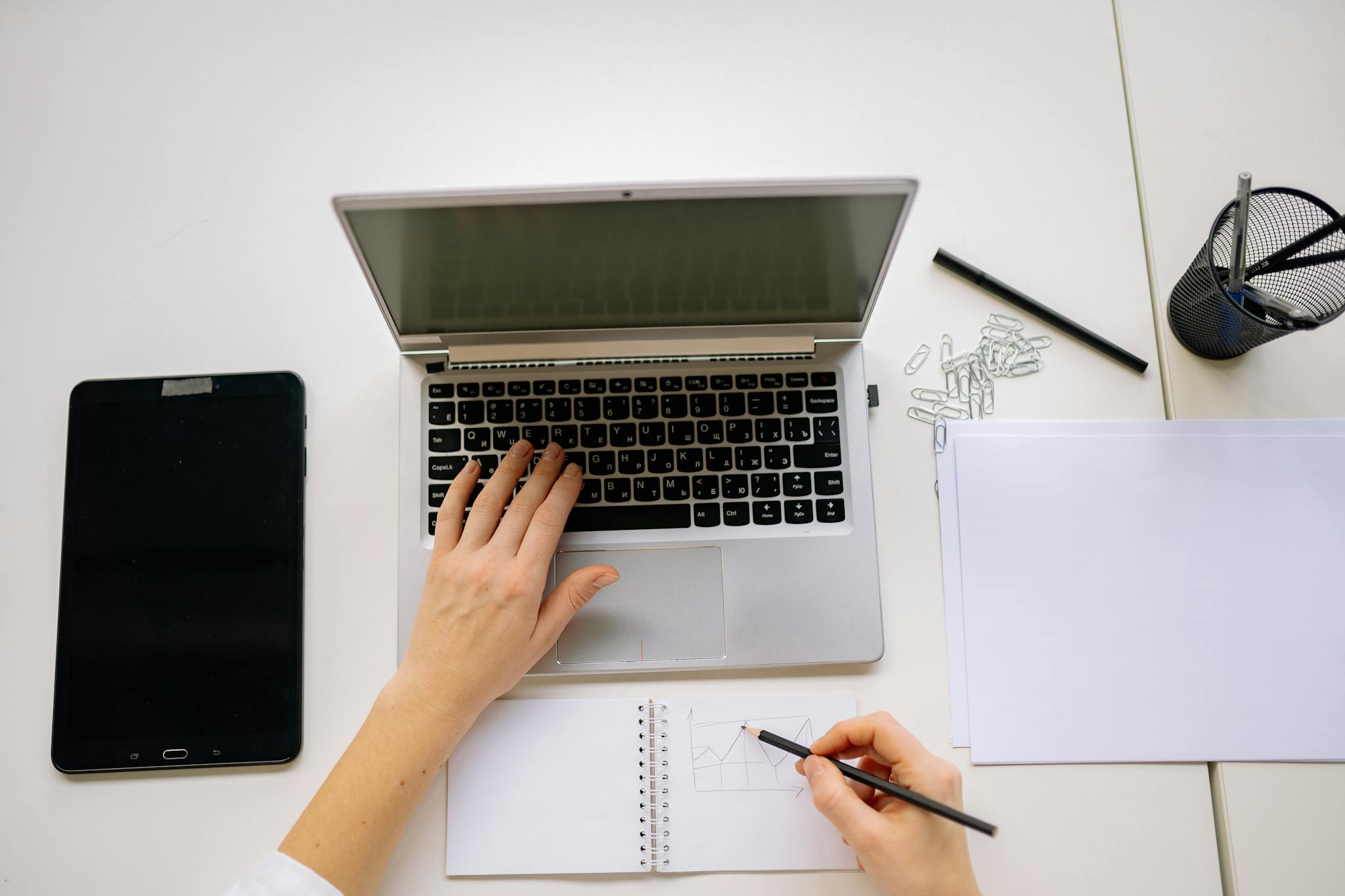 Hands multitasking on laptop and notepad with tablet on desk, perfect for productivity themes.